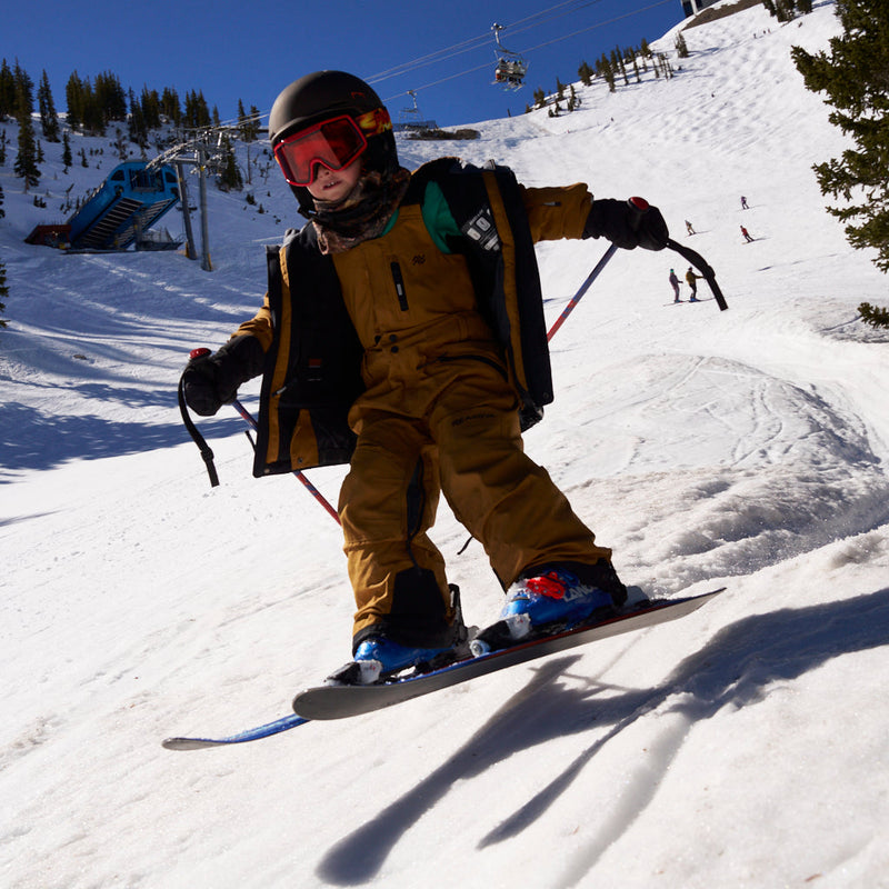 | oliver_rips.JPG | A child skier wearing a tan snowsuit, black helmet, and red goggles is skiing down a snowy mountain slope with ski poles and a ski board visible.