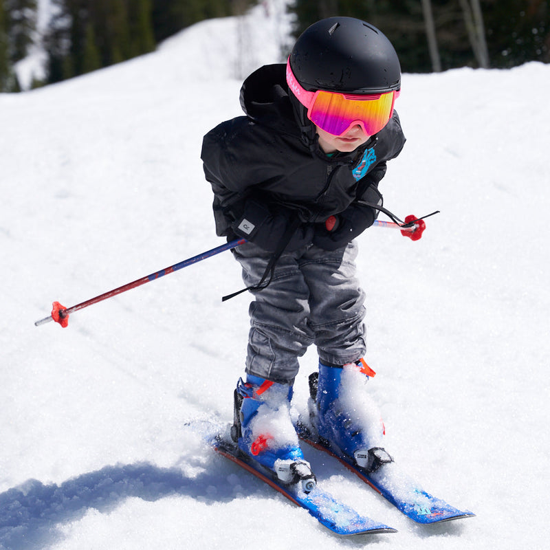 | ripper.JPG | A child skier wearing a black jacket, gray ski pants, blue ski boots, a black helmet, and vibrant pink/orange goggles, leaning forward on blue skis with ski poles in a snowy outdoor setting.
