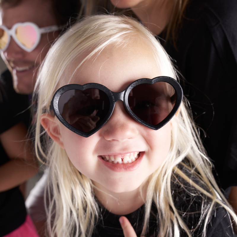 Smoke | photo_of_person.JPG | A close-up lifestyle shot of a young girl with blonde hair wearing black heart-shaped sunglasses, with other people partially visible in the background.