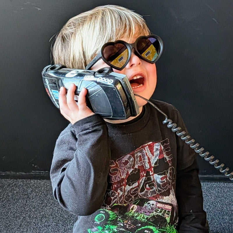 Smoke | photo_of_boy.JPG | A young child sitting on a dark carpeted floor, holding a toy walkie-talkie to their ear as if talking on a real phone. The child is wearing round black sunglasses, a dark gray graphic long-sleeve shirt with green monster truck graphics, and blue jeans.